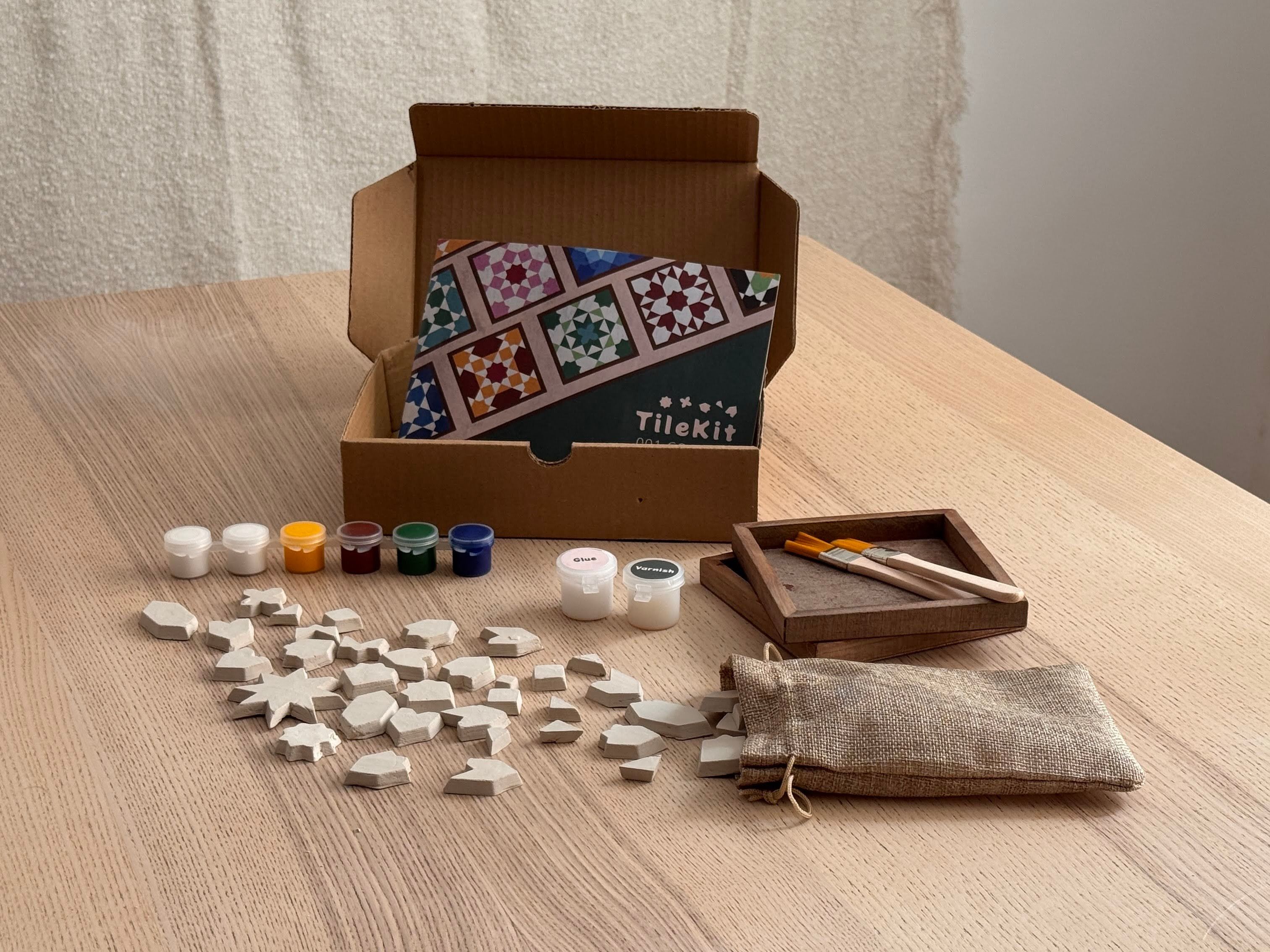 Capsa de TileKit oberta sobre una taula amb peces d’argila, pintures, pinzells i bases